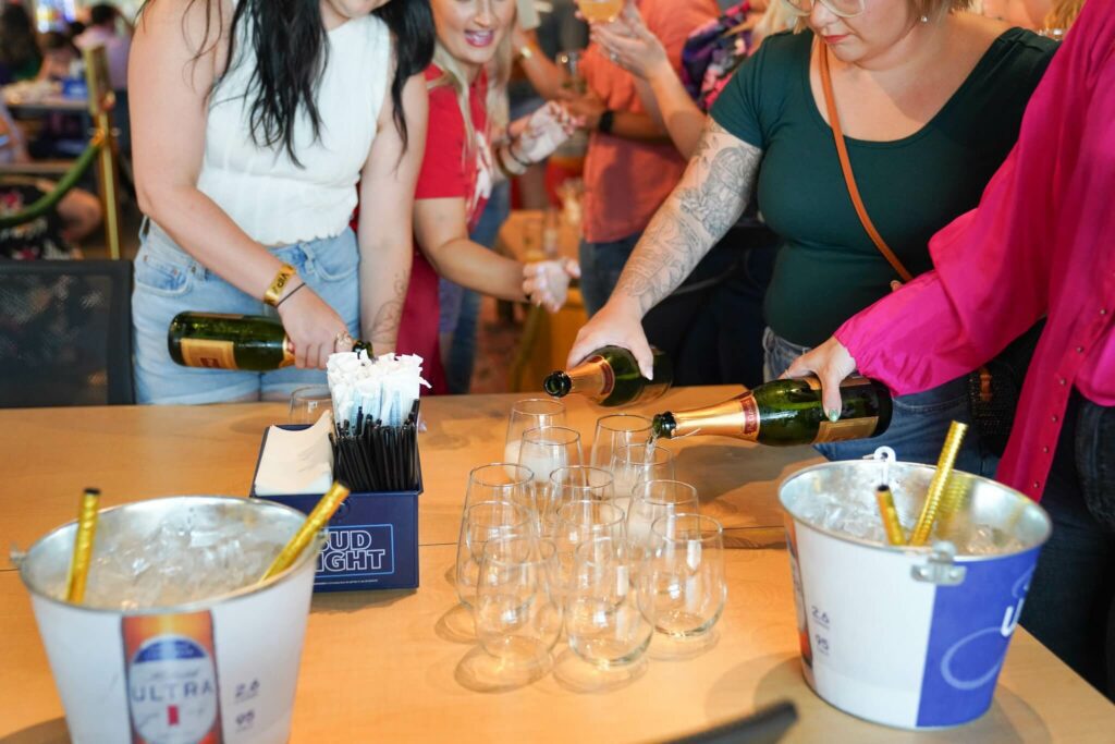 A group of people, mostly women, standing around a table pouring drinks and socializing in a lively, festive environment with colorful decor and lighting.