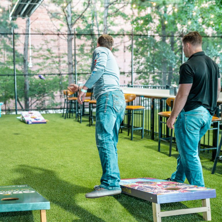 An image depicting an outdoor seating area with people playing cornhole and other lawn games, surrounded by greenery and a lively atmosphere, suggesting a fun and social environment.